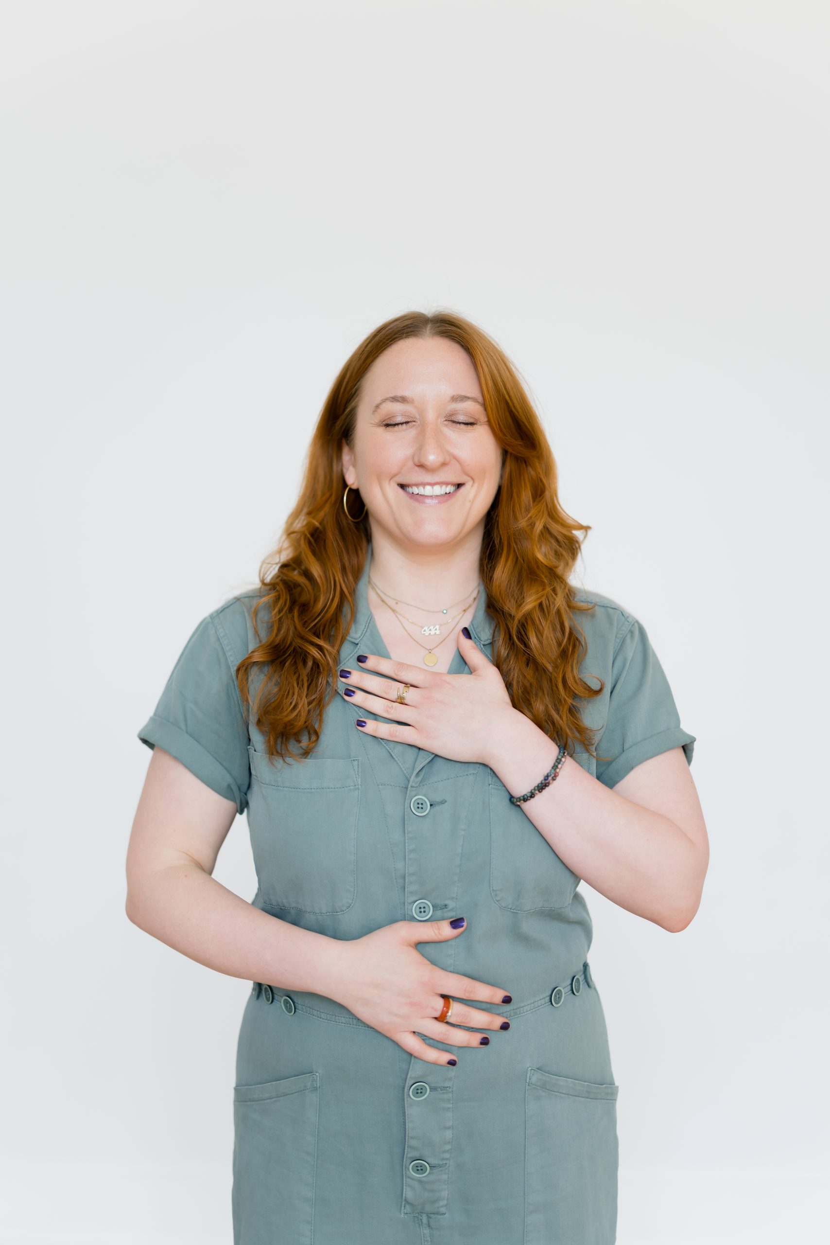 Katie Healy in a sage jumpsuit demonstrating a breathing technique with one hand on her belly, the other on her heart
